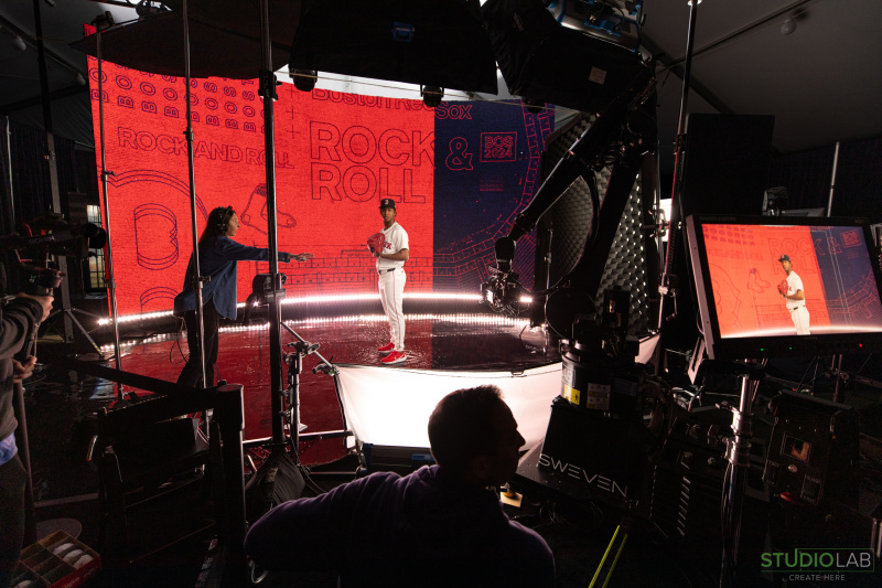 A photo of a studio with a player standing in the middle posing for the camera while another person is giving directions.