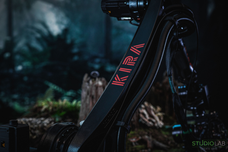 Close-up shot of a camera rig labeled "KIRA" with cables attached, set against a dark, forest-like background, hinting at a cinematic or filming setup.