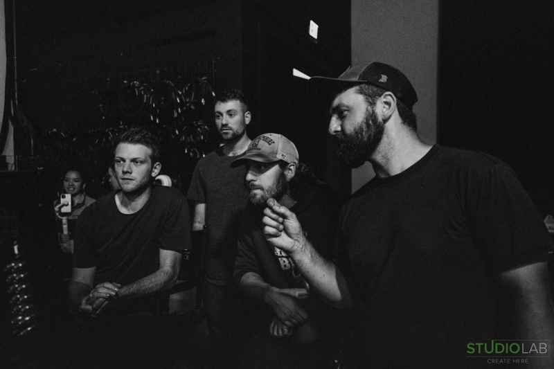 A group of 4 people huddle together in a dark room, focusing intently on something off-camera. One person gestures as if explaining something out.