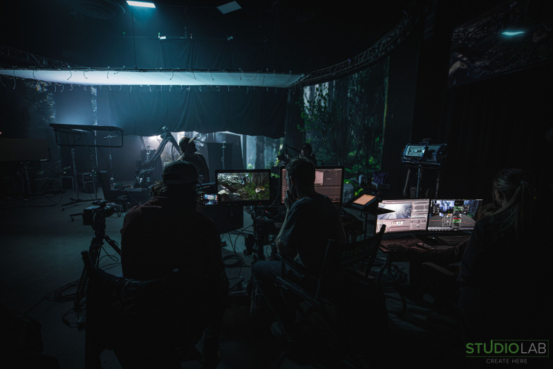 A dark set shows a team of operators in front of multiple screens, reviewing footage while a camera crew captures scenes under soft lighting in the background.