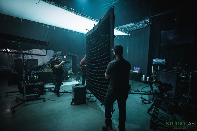A camera operator is filming on a dark set while two crew members adjust a large curved lighting screen. Equipment and cables are scattered across the floor.