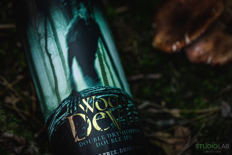 Close-up of a beer can labeled "Wood Devil Double Dry Hopped Double IPA," lying on forest floor. The can’s eerie design features a dark, shadowy figure.