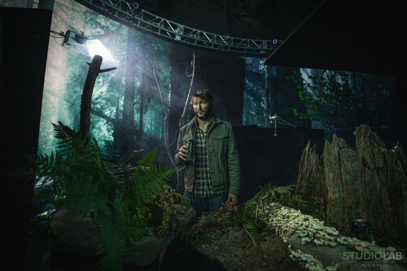 A man stands in a forest-like set holding a beer can, surrounded by ferns and natural elements. Creating a dark woods setting for a commercail.