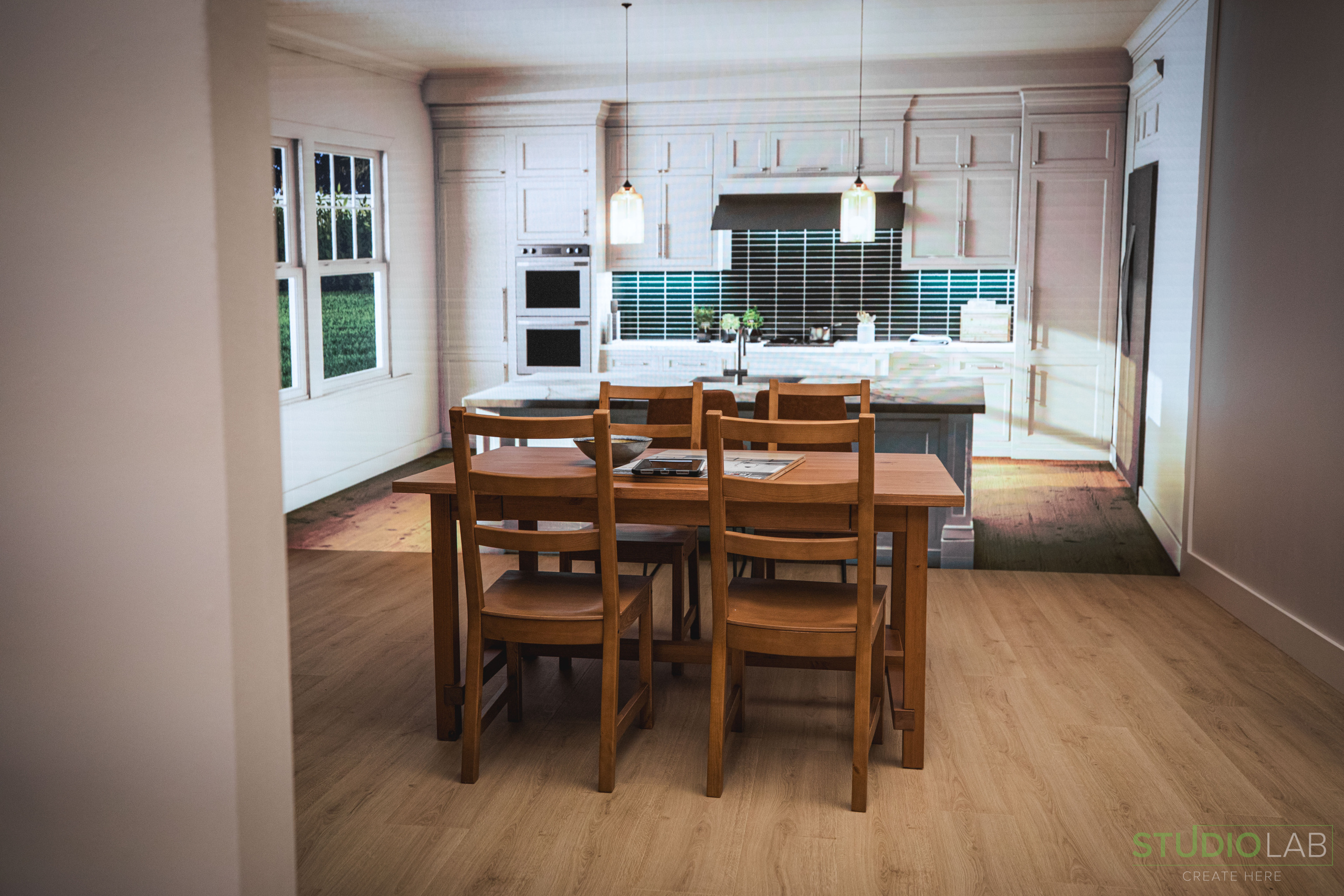 A simple wooden dining table and chairs are placed in front of a projected backdrop of a modern kitchen with white cabinetry.