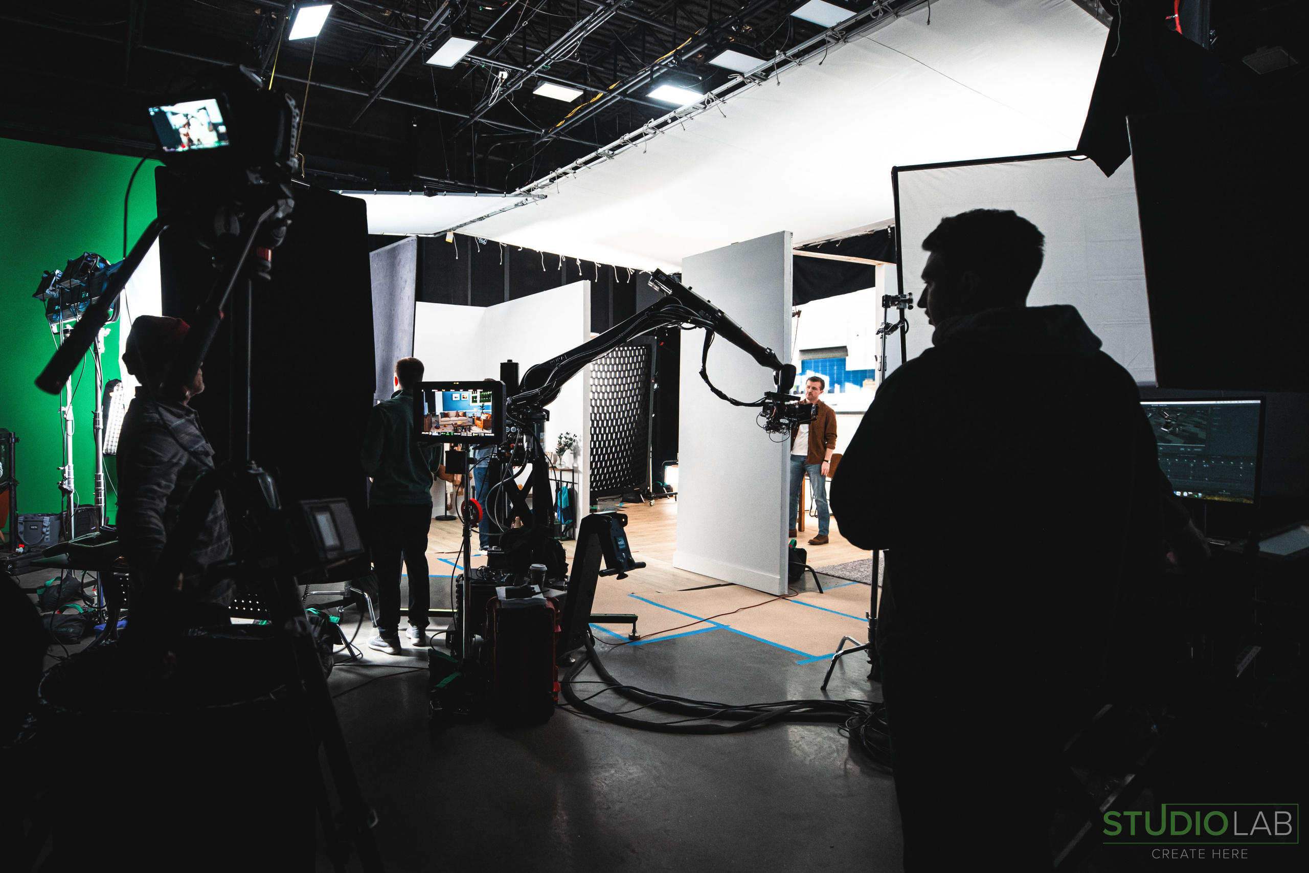 A behind-the-scenes view of a production set with lights, camera equipment, and a crew member silhouetted against a brightly lit stage.