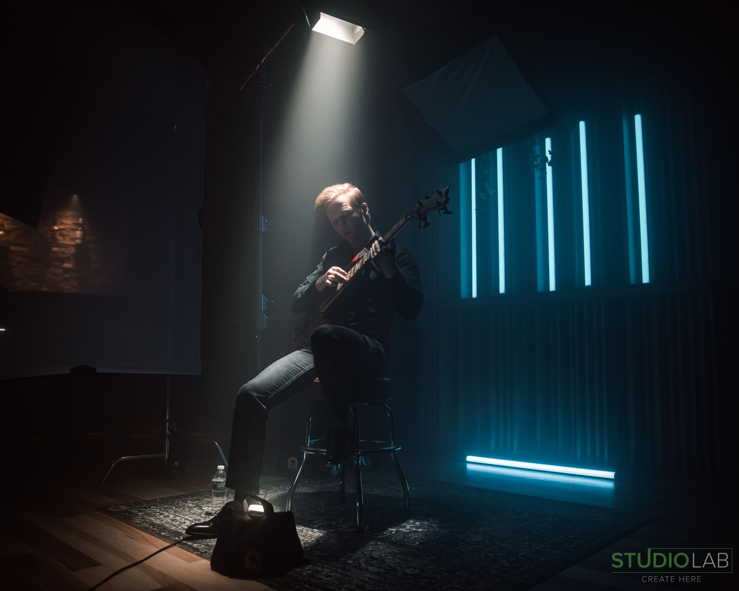 A musician sits on a stool, illuminated by a spotlight, playing a guitar in a dimly lit studio with vertical neon lights in the background.