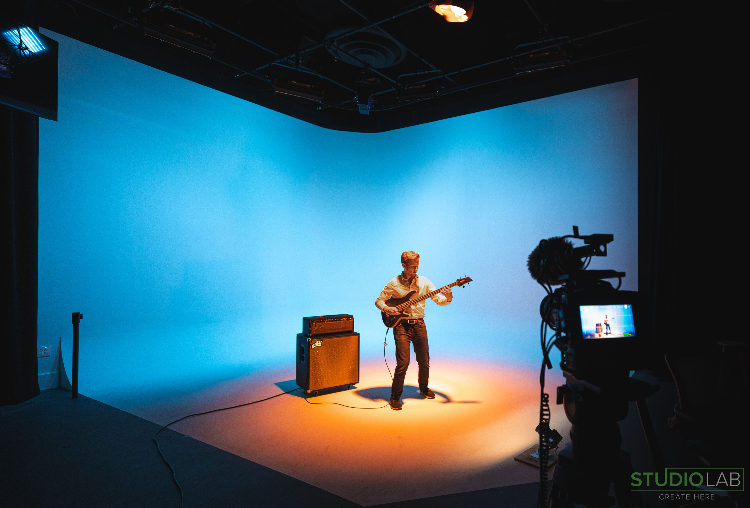 A musician plays a guitar on a brightly lit stage with a gradient blue-to-orange backdrop, while a camera captures the performance.