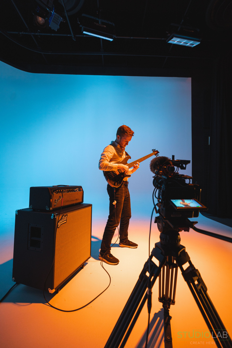 Musician playing a guitar on a brightly lit set with a gradient blue-to-orange backdrop. A camera on a tripod captures the performance.