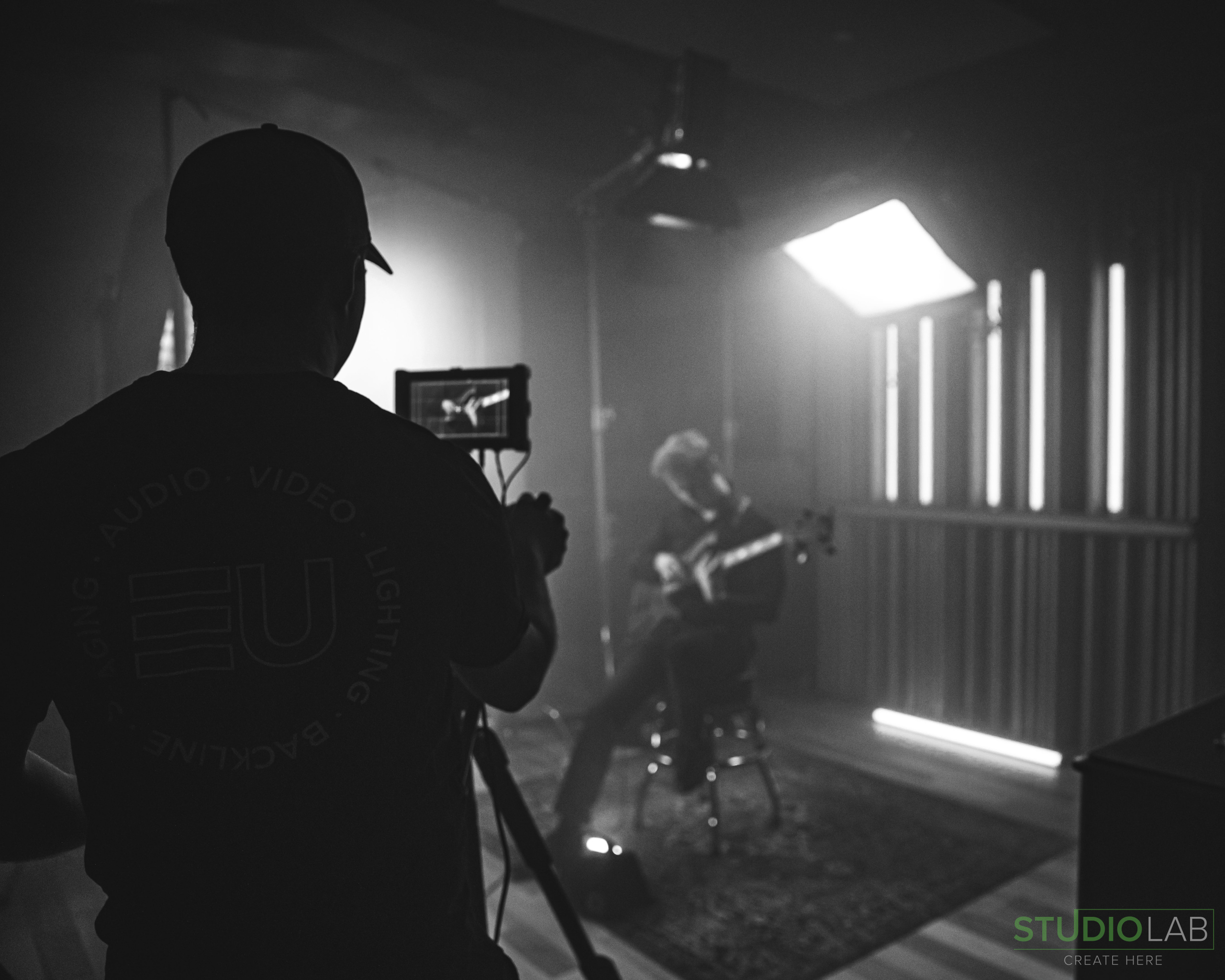 Black-and-white shot of a musician playing guitar in a moody, dimly lit studio while a cameraman films the scene, creating a dramatic atmosphere.