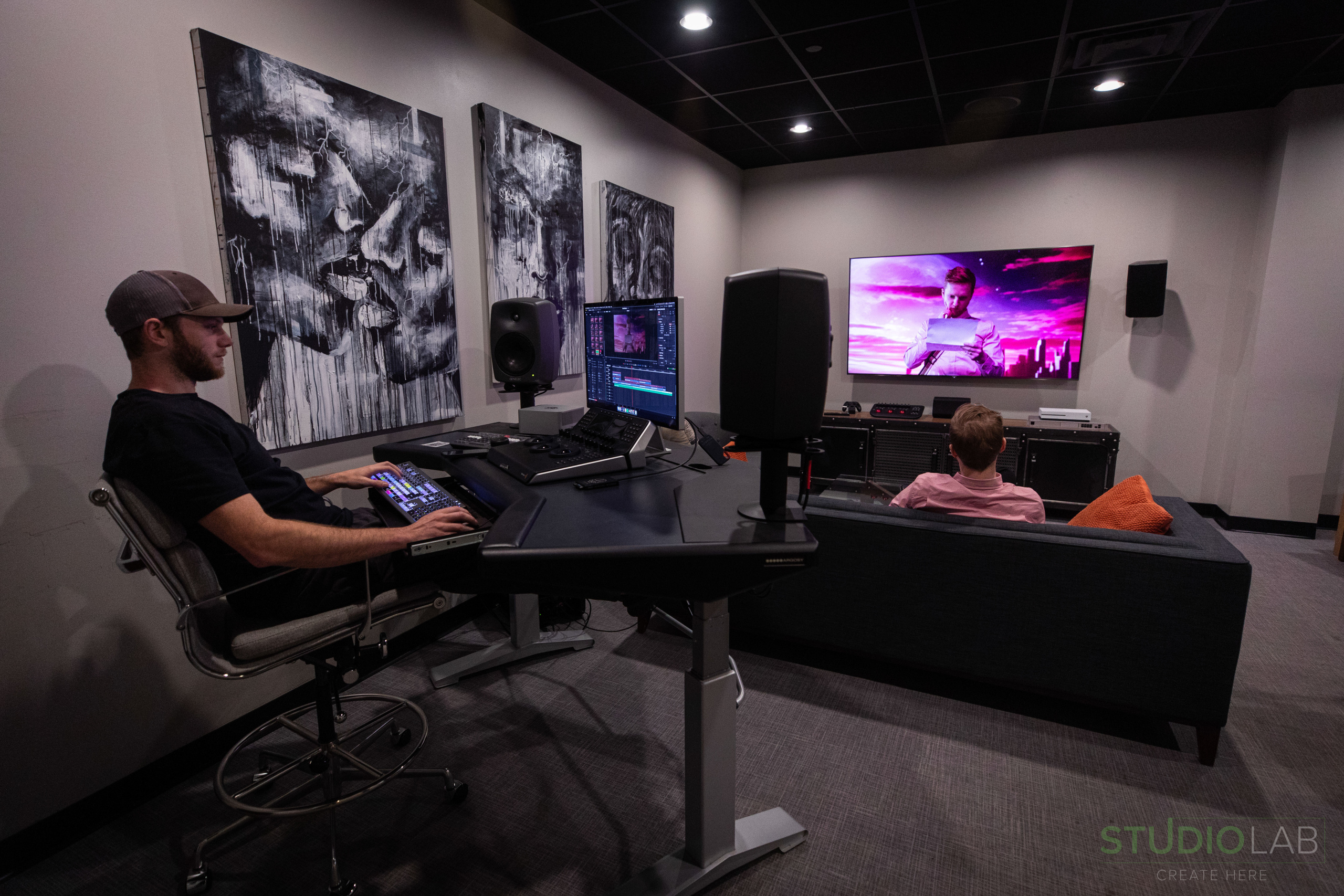 Editing suite with a person at a control desk, another on a couch watching a large screen, and abstract black-and-white artwork on the wall.