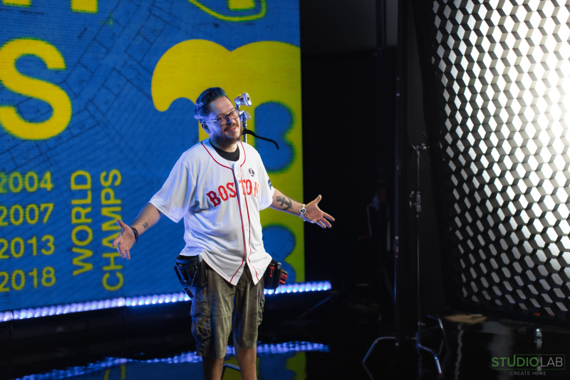 A person in a Boston Red Sox jersey stands on set, smiling with arms open. Behind them is a large LED screen displaying "World Champs" and the championship years.