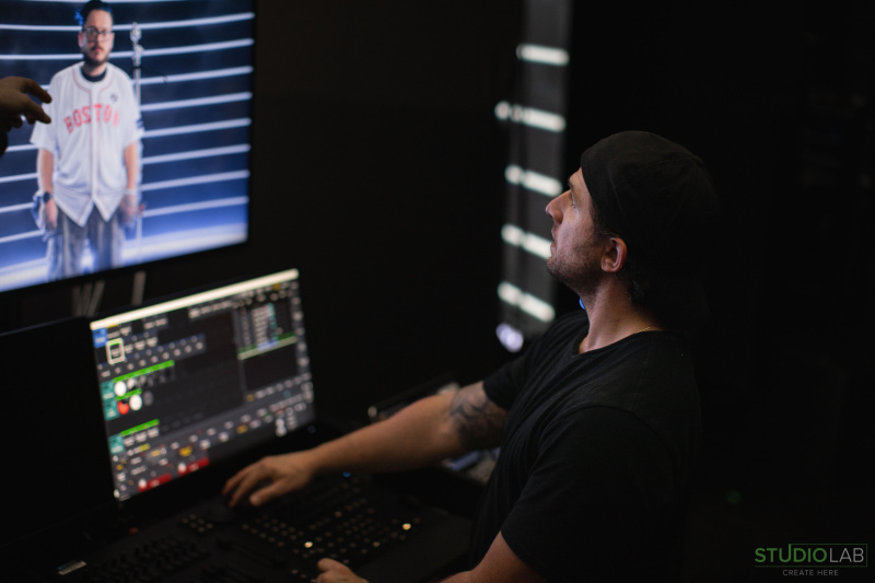 A technician in a black shirt and backwards cap sits at a control desk, focused on a screen displaying a person wearing a Boston Red Sox jersey in a studio.