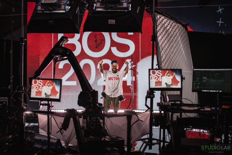 A man stands in front of a large LED screen displaying "BOS 2024" with thumbs up, surrounded by cameras and screens displaying his image.