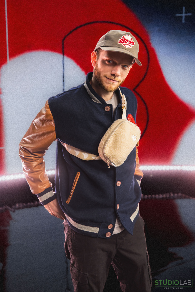 A man wearing a Red Sox cap, letterman jacket with brown sleeves, and a beige cross-body pouch, posing confidently in front of a Red Sox backdrop.