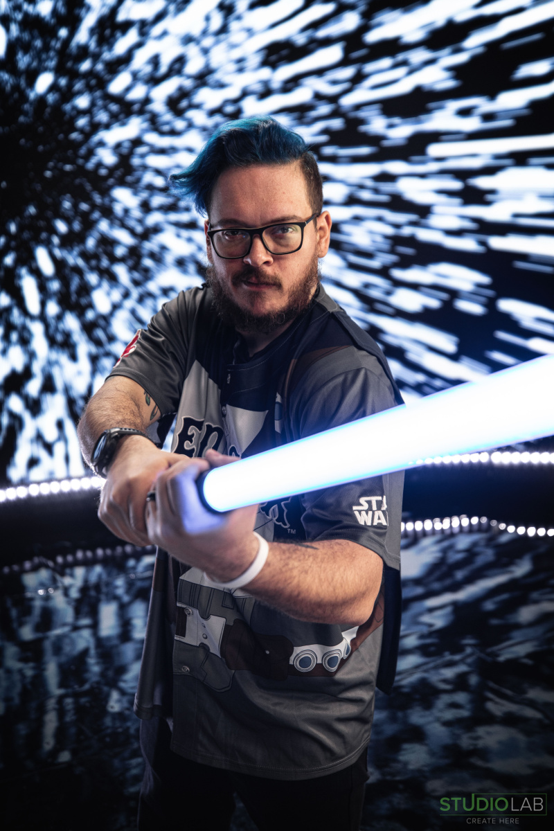 A man with blue hair and glasses holding a glowing lightsaber prop, with a Star Wars-style hyperspace background behind him.