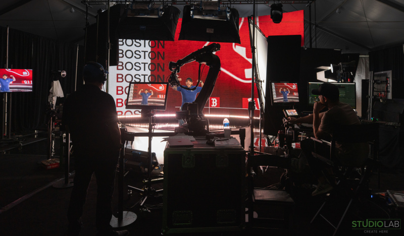 A production stage with a robotic camera, lights, and crew filming a subject in front of a Boston-themed LED backdrop.