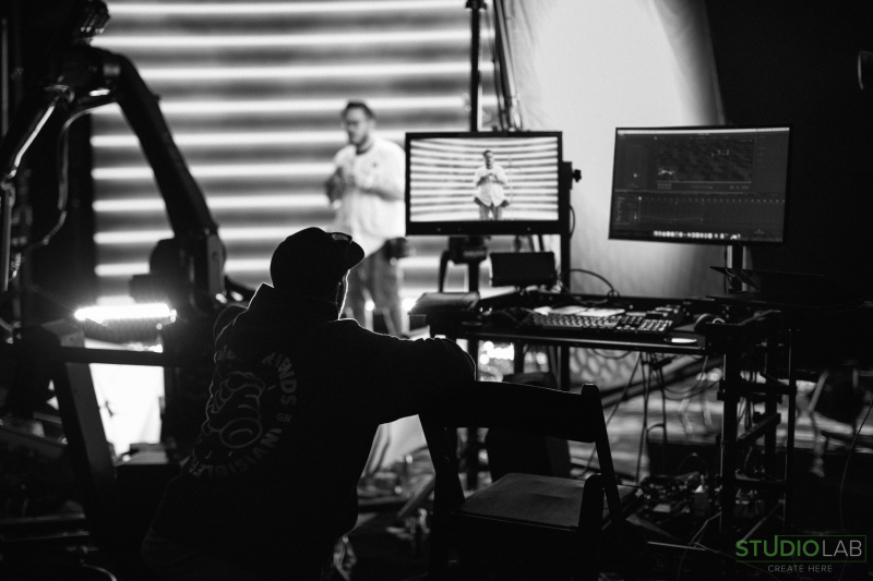 Black-and-white shot of a production crew monitoring a screen during filming. The subject in a Boston jersey is visible on the screen and stage, surrounded by equipment.