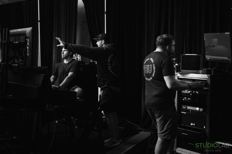 A black-and-white image of the production team working. One person gestures toward a screen while another operates equipment under dark curtains.