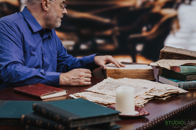 Man in a blue shirt is sitting at a wooden desk covered with old books, manuscripts, and a lit candle, he examines a large, ancient book.