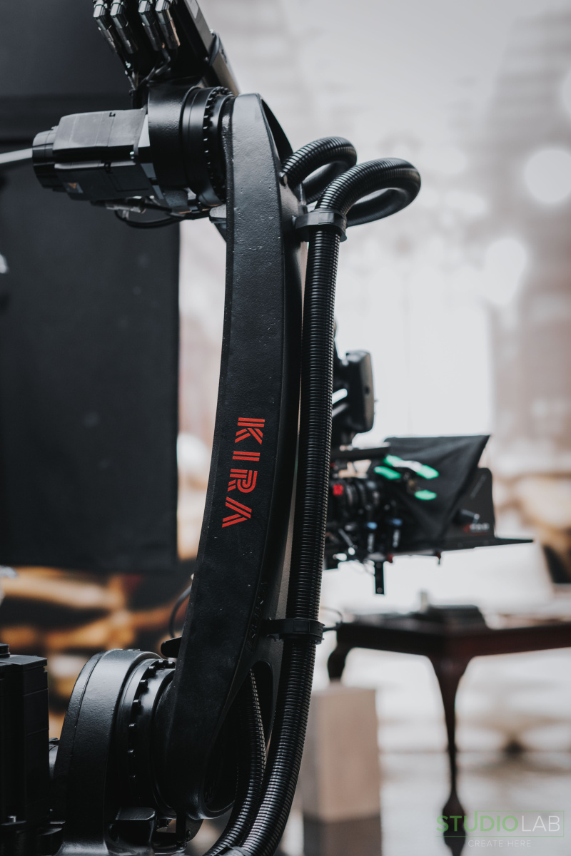 A close-up of a KIRA robotic camera arm on set, with the camera and part of the set visible in the background.