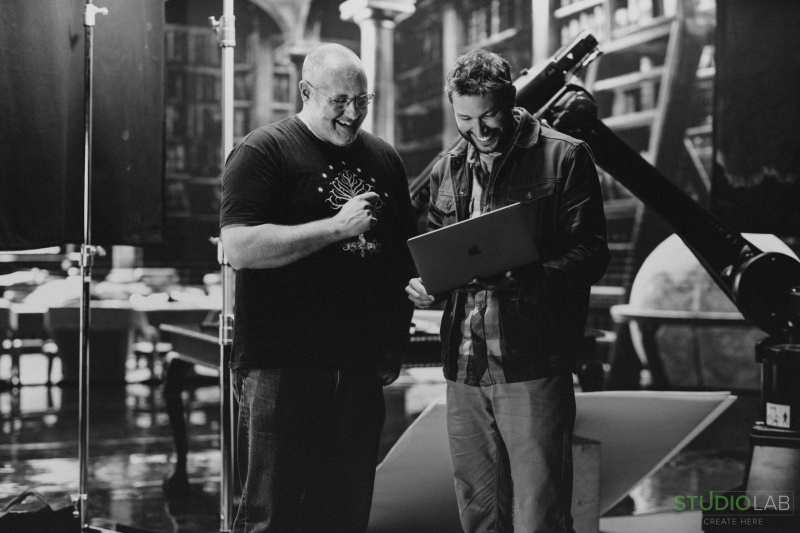 Two men smiling and reviewing content on a laptop on set, with a library backdrop and a robotic camera arm visible in the background.