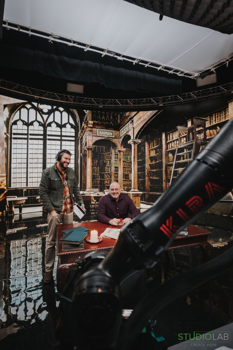 A director and actor on set with a library backdrop, the actor seated at a wooden desk, lit by a candle, with a robotic camera arm in the foreground.