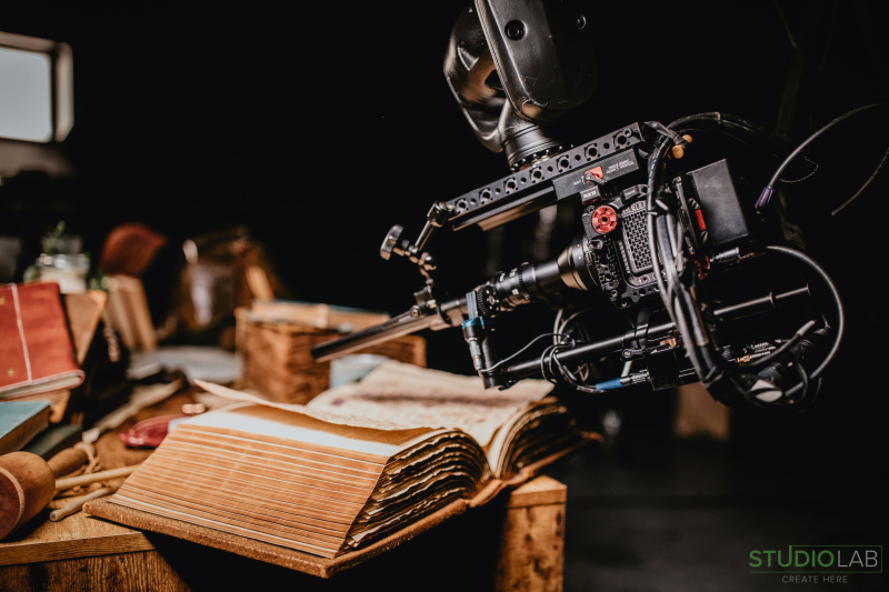 Close-up of a robotic arm labeled KIRA, positioned to film an open book surrounded by old manuscripts, leather-bound books, and other props.