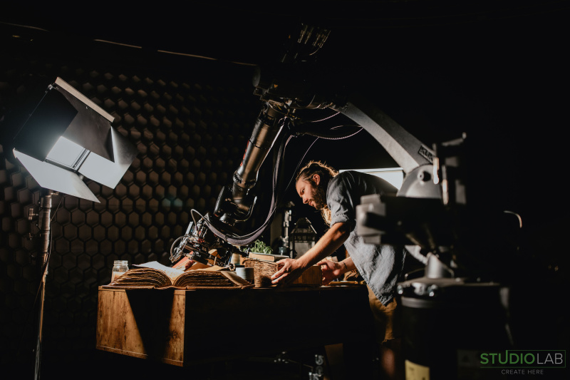 Cinematic setup with a man adjusting props on a table under focused lighting, KIRA robotic arm poised over open books, enhancing a medieval scene.