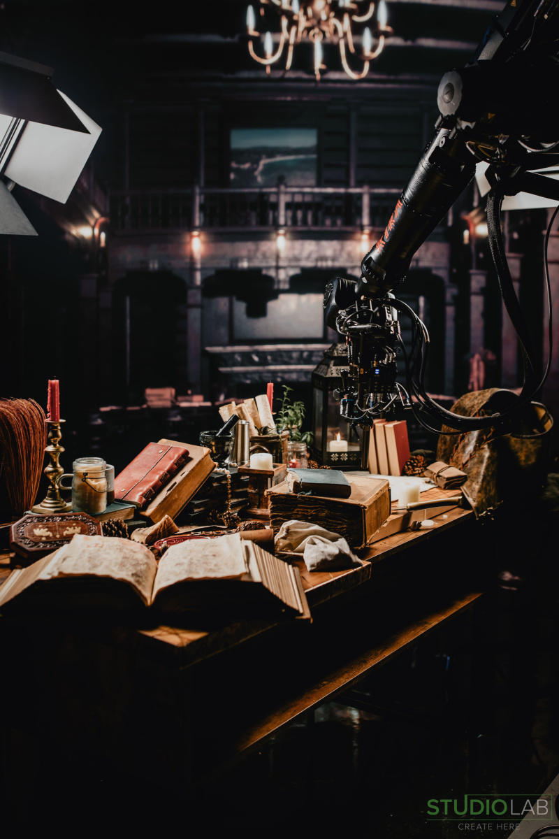 A dimly lit scene with a medieval table setup of books, candles, and scrolls, as a robotic camera arm hovers, capturing the setup.