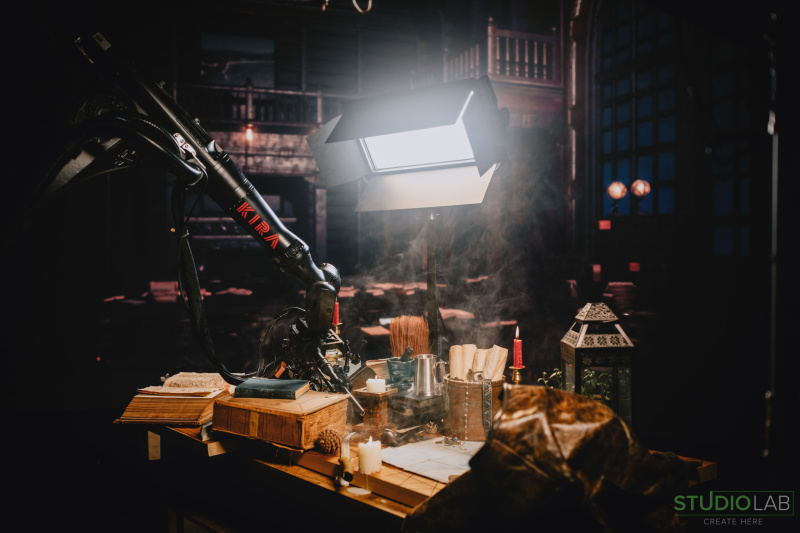 A production setup featuring the KIRA robot arm filming a medieval-themed desk with candles, books, and various props under dramatic lighting.