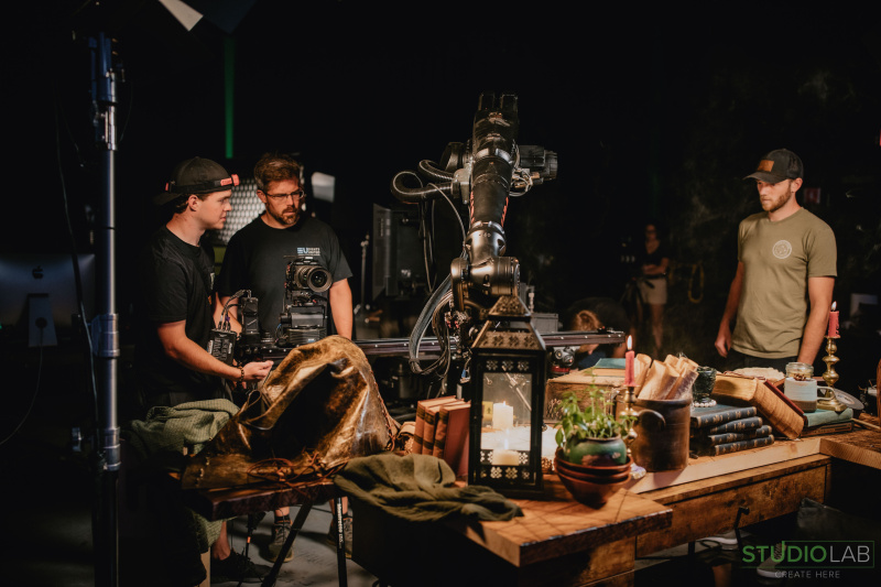 Three crew members on a set, one operating a camera, with a large robotic arm positioned over a table filled with books, candles, and props.