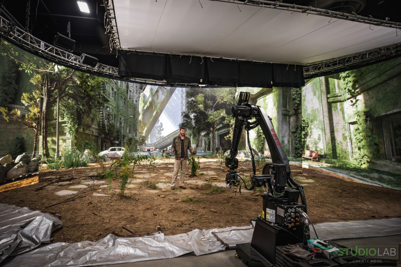 A camera rig captures a scene featuring an actor in a post-apocalyptic setting, surrounded by overgrown vegetation, abandoned cars, and crumbling buildings.