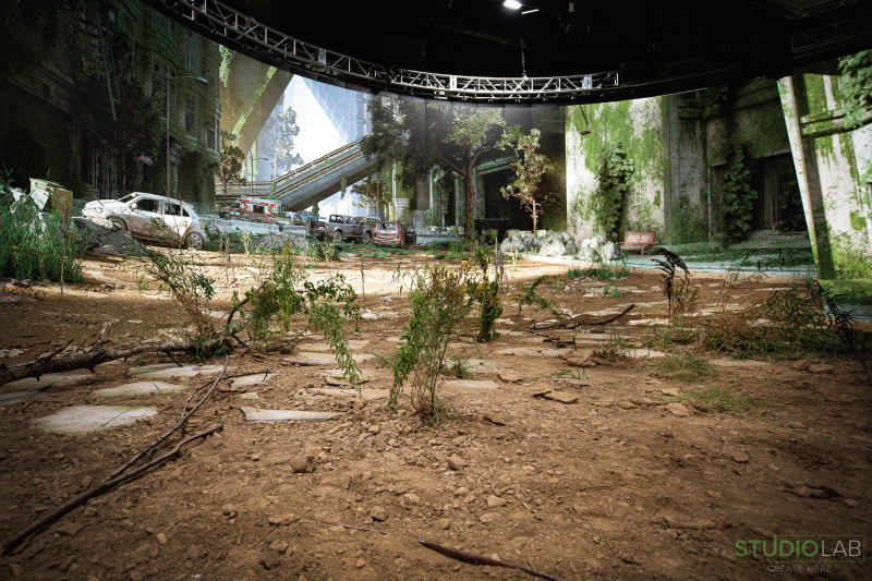 A detailed set portrays a post-apocalyptic cityscape with overgrown plants, abandoned vehicles, and crumbling buildings. Dirt and debris cover the ground.