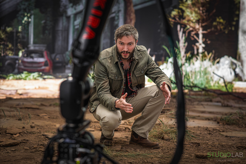 A man crouches intently on a dirt-covered set, dressed in outdoor gear, with a large camera rig in the foreground. The background depicts an overgrown, abandoned scene.