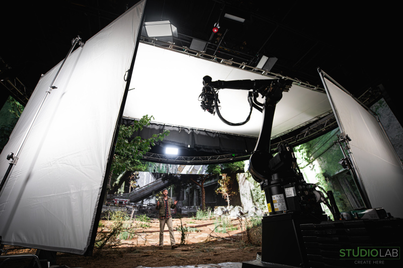 A man stands on a post-apocalyptic set with robotic cameras surrounding him. Overhead lighting panels and greenery create an immersive outdoor environment.