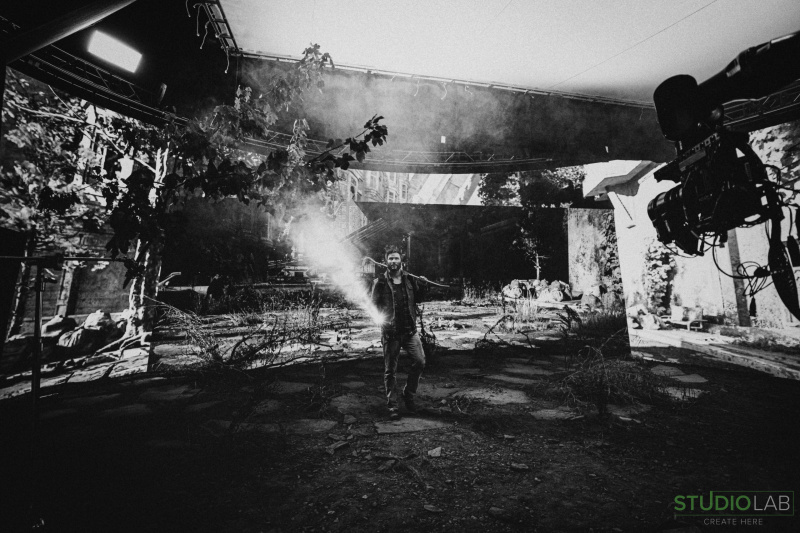 A man stands in a decaying urban environment, holding a flashlight while surrounded by overgrown foliage and debris. A camera rig is visible on the side.