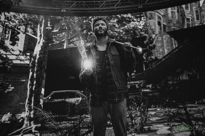 A man stands in a post-apocalyptic setting, holding a flashlight in one hand and a weapon over his shoulder, surrounded by ruined buildings and overgrown vegetation.
