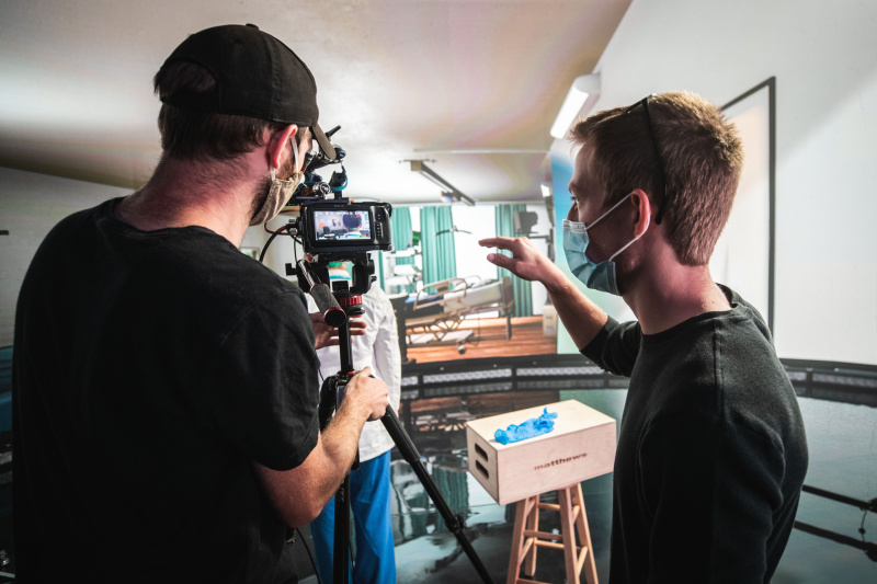Two crew members, wearing masks, film a scene on a hospital set. One operates the camera while the other directs the scene.