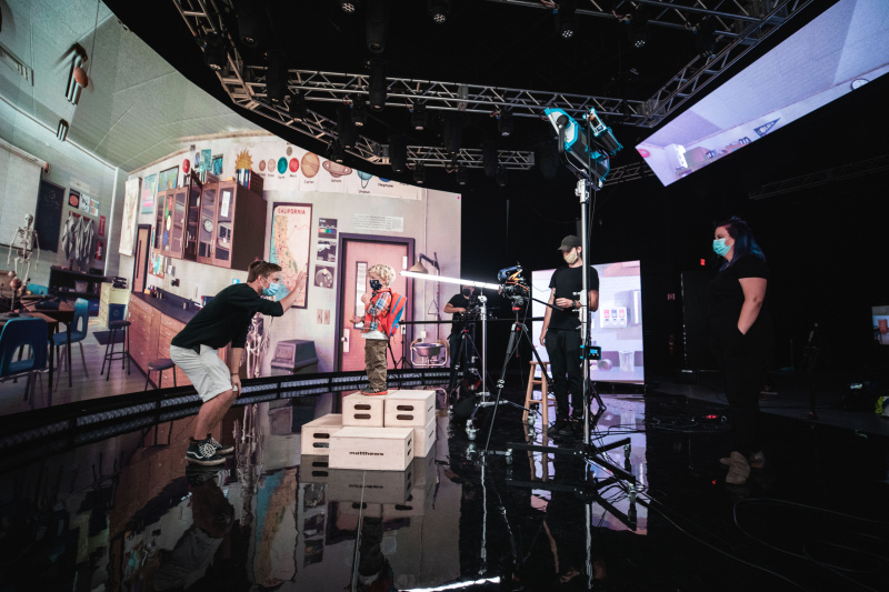 A film crew directs a young child on a set designed as a classroom. The crew and child wear masks, with lights and cameras positioned around them.