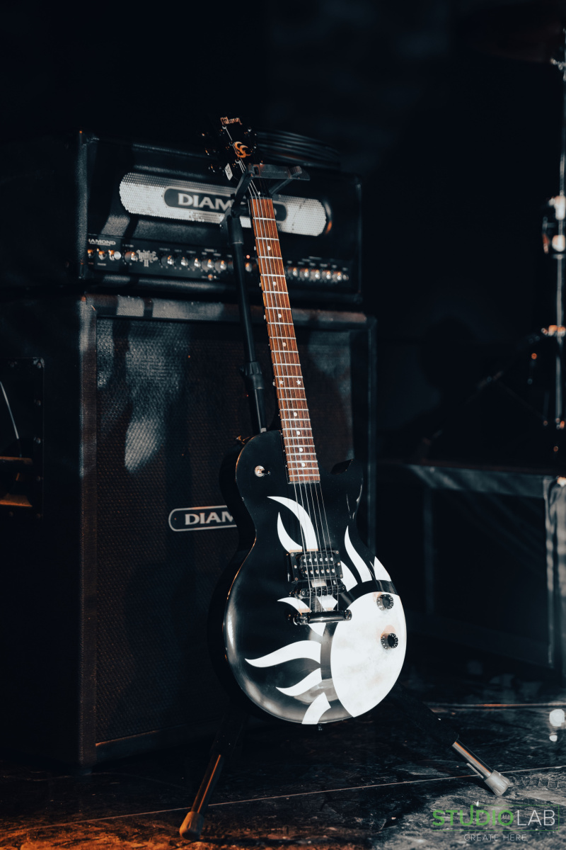A black electric guitar with a white tribal sun design rests against a Diamond brand amplifier on a dimly lit stage, ready for a rock performance.