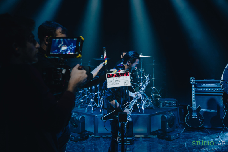 A cameraman captures footage of a rock guitarist during a live performance on stage, with drums and other musical equipment visible in the background.