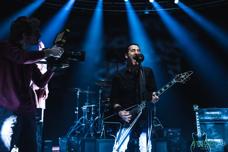 A lead guitarist passionately plays his guitar on stage under bright blue lights, with a camera operator capturing the moment, during a live performance.