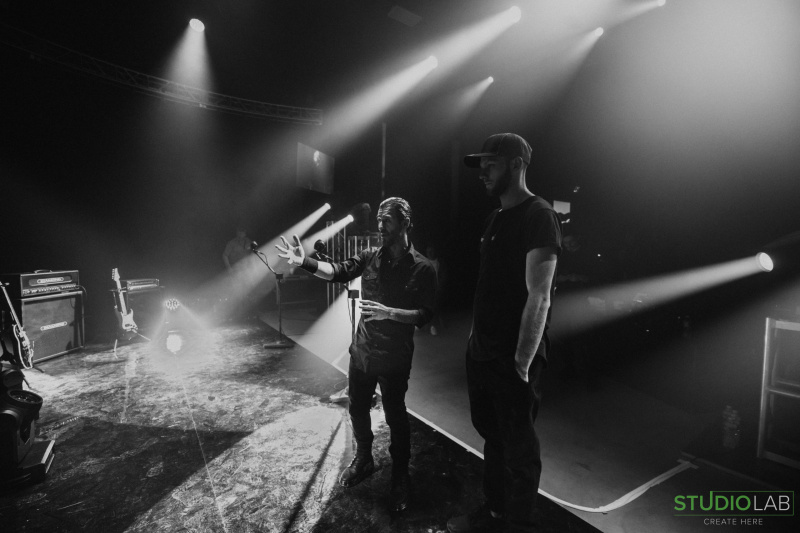 The band Godsmack, standing on a stage drenched in light beams, prepares for a performance while discussing stage direction with the lighting crew.