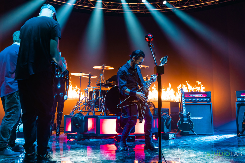 A guitarist on stage in a dark setting with spotlights, playing passionately as flames burn behind the drum set. Cameramen in black shirts capture the moment.