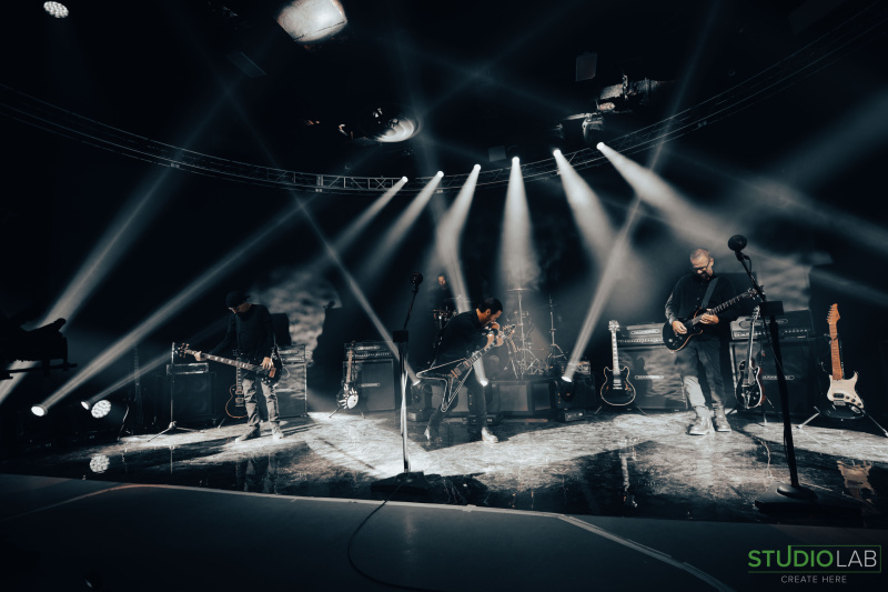 A rock band performs on stage, with light beams. The lead guitarist kneels while other band members play their instruments, creating an intense atmosphere.