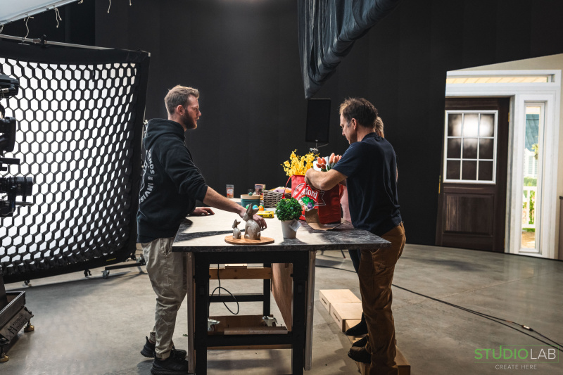 On set, two men set up a kitchen scene for filming, with a Hannaford grocery bag on the table and props like flowers and ceramic bunnies placed nearby.