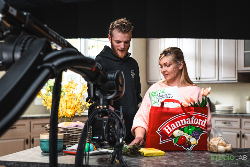 A camera films two people in a kitchen setting. The woman holds a red Hannaford grocery bag filled with products, while the man observes beside her.