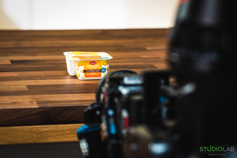A camera focuses on a container of Hood Medleys cottage cheese with pineapple added. The product sits on a wooden surface, ready to be captured on film.