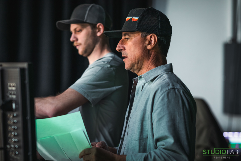 Two crew members focus intently while working on set. One holds a green sheet of paper, as they collaborate on the project.