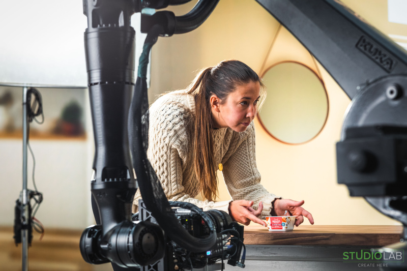 A woman in a cream-colored sweater leans over a wooden table, carefully arranging a Hood Medleys cottage cheese container while a robotic camera rig captures the scene.